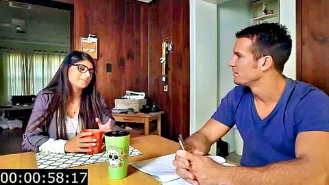Clothed brunette with glasses chats intimately with muscular man at home table holding skull mugs