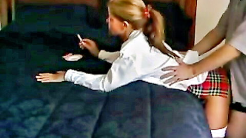 Blonde teen in schoolgirl skirt on bed with lollipop, hand on back